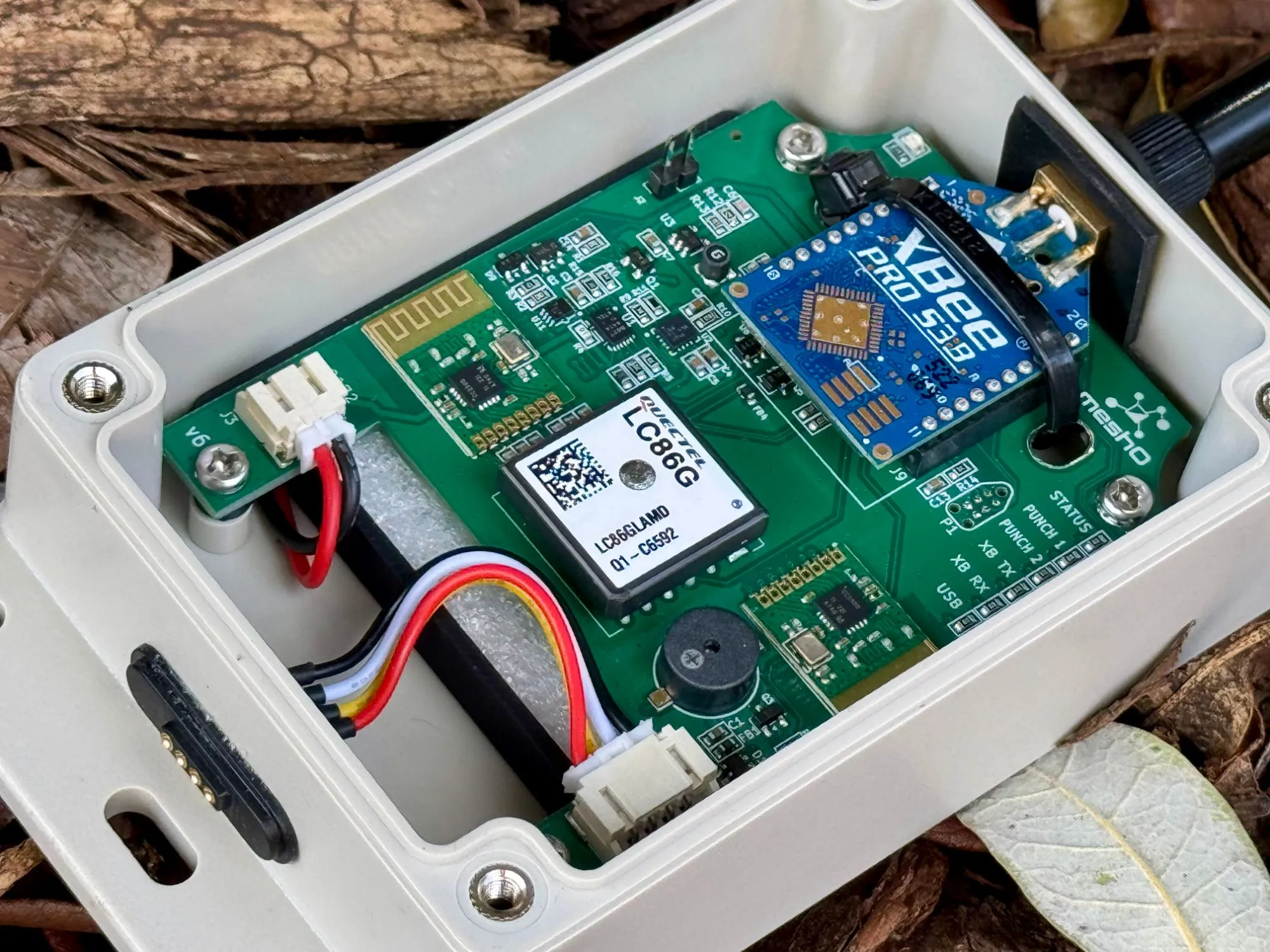 A meshO Control with its lid off, lying on bark: the green v6 circuit board with a Quectel LC86G GPS module in the middle, a blue XBee PRO S3B mesh radio top right, two small 2.4 GHz radio boards top left and bottom right, a buzzer and a row of status lights.