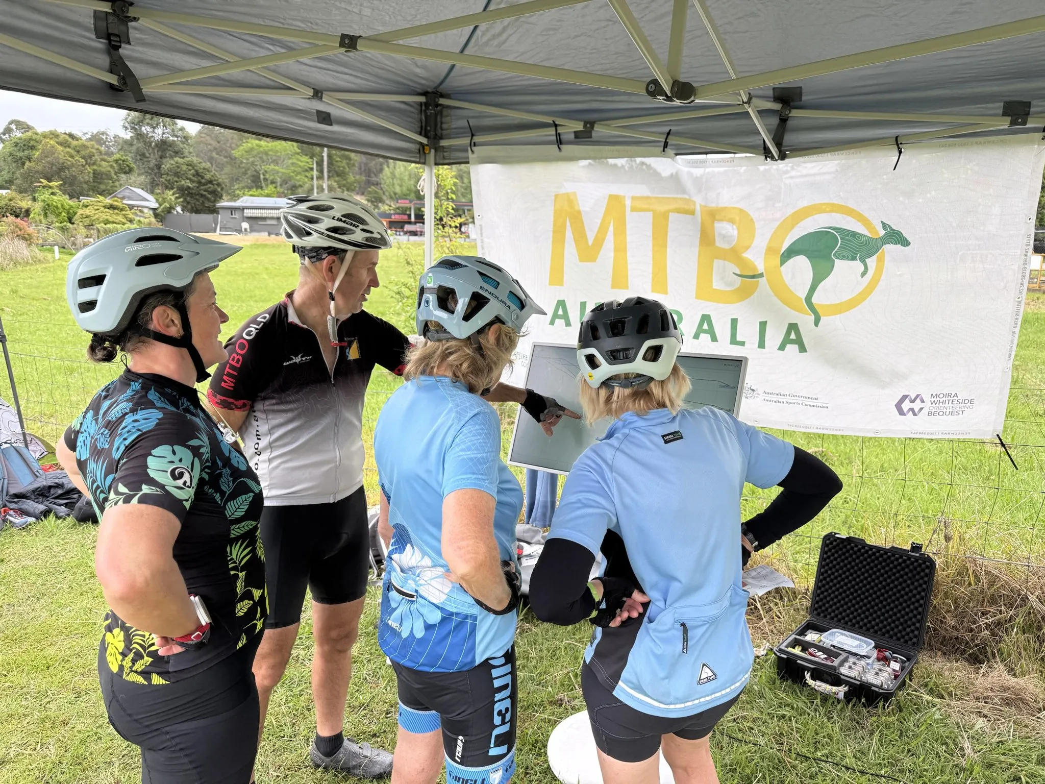 Four riders in helmets and cycling jerseys gather round a screen under the MTBO Australia banner, one pointing at a route on it