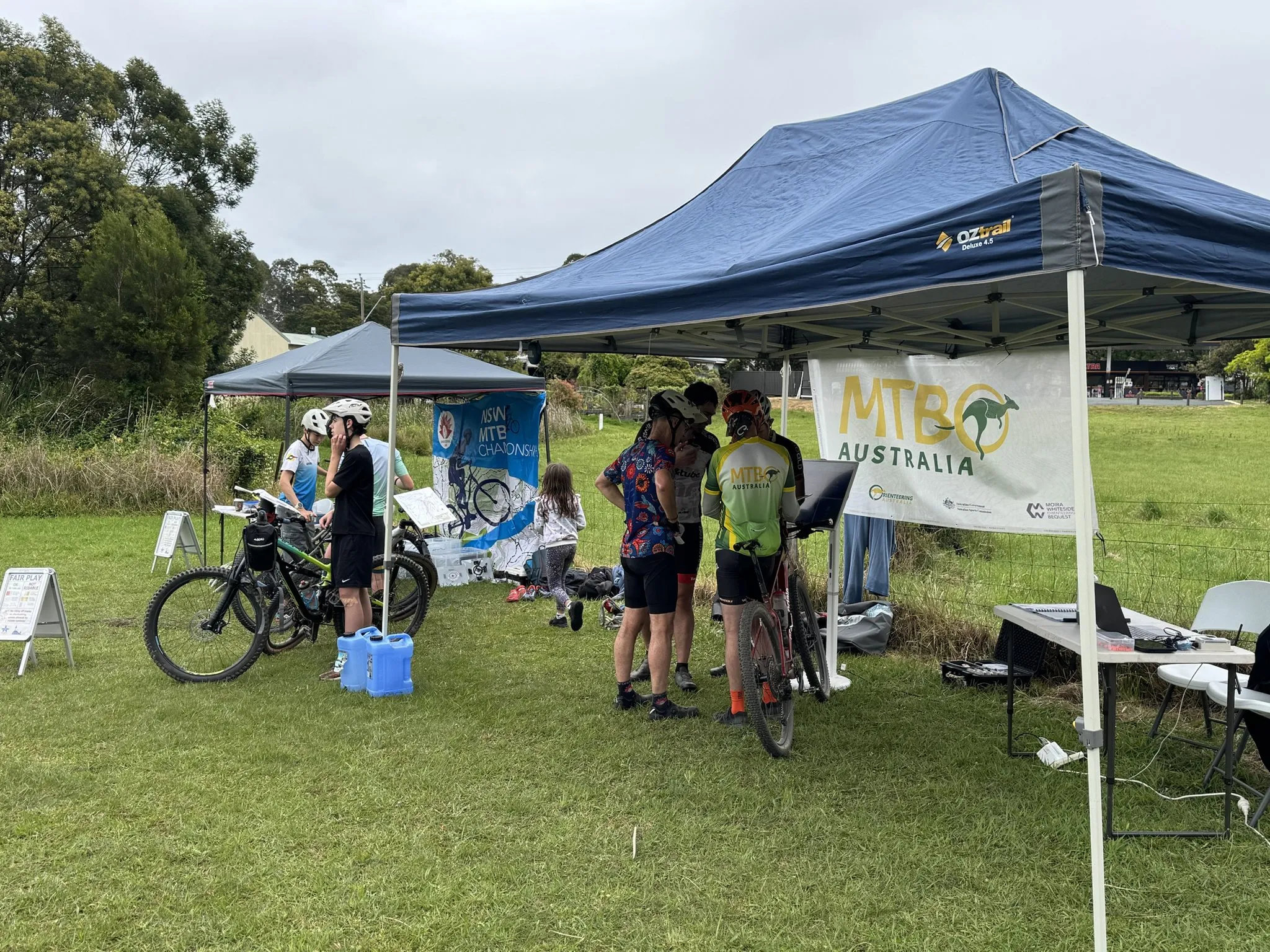 Riders with their bikes gathered at a screen under the blue MTBO Australia marquee, with the NSW MTBO Championships tent behind