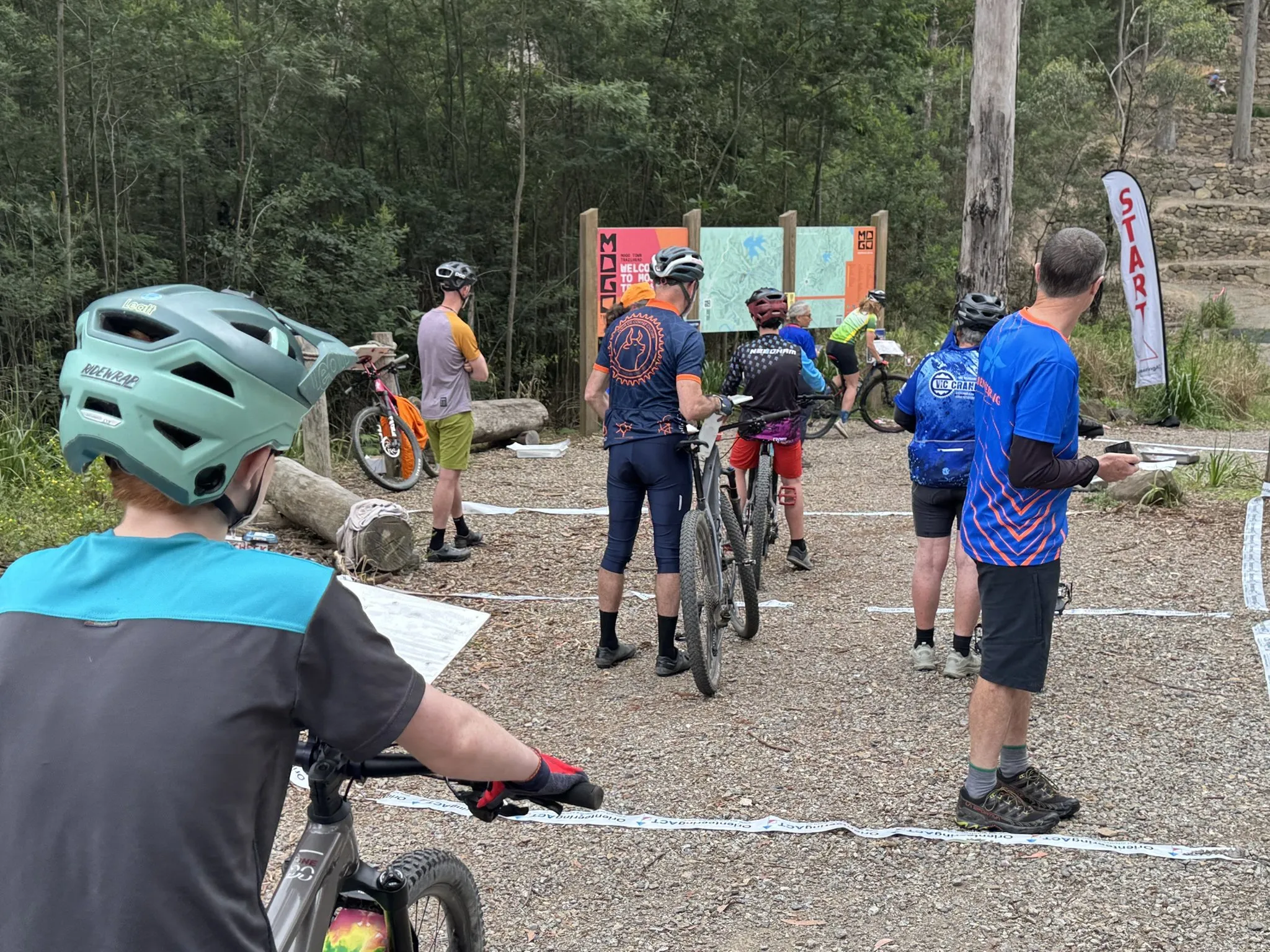 Riders with their bikes waiting in taped start lanes in front of the Mogo Trails welcome sign and a red START flag