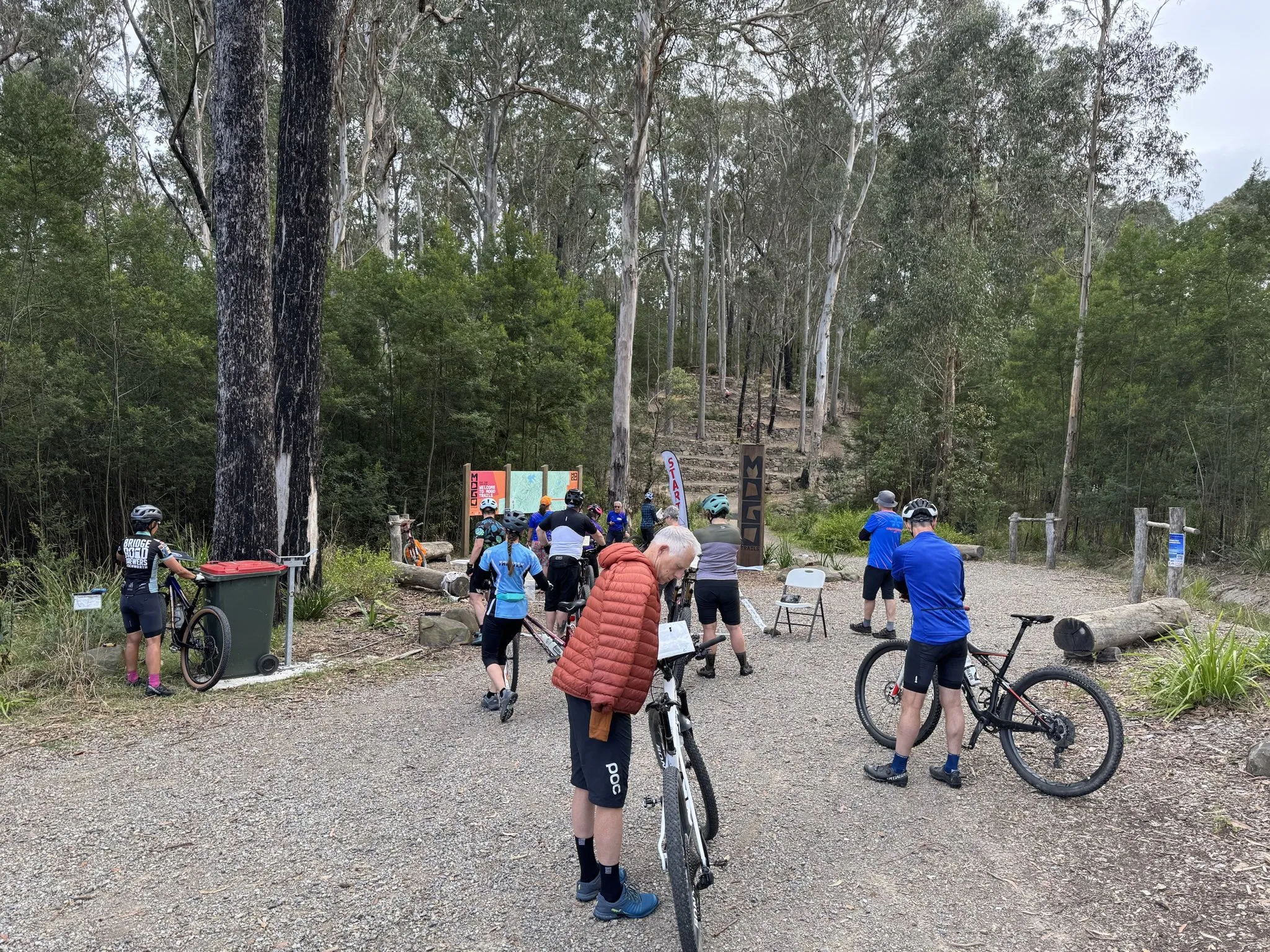 Riders and bikes in the gravel start area under tall gum trees at the Mogo Trails trailhead