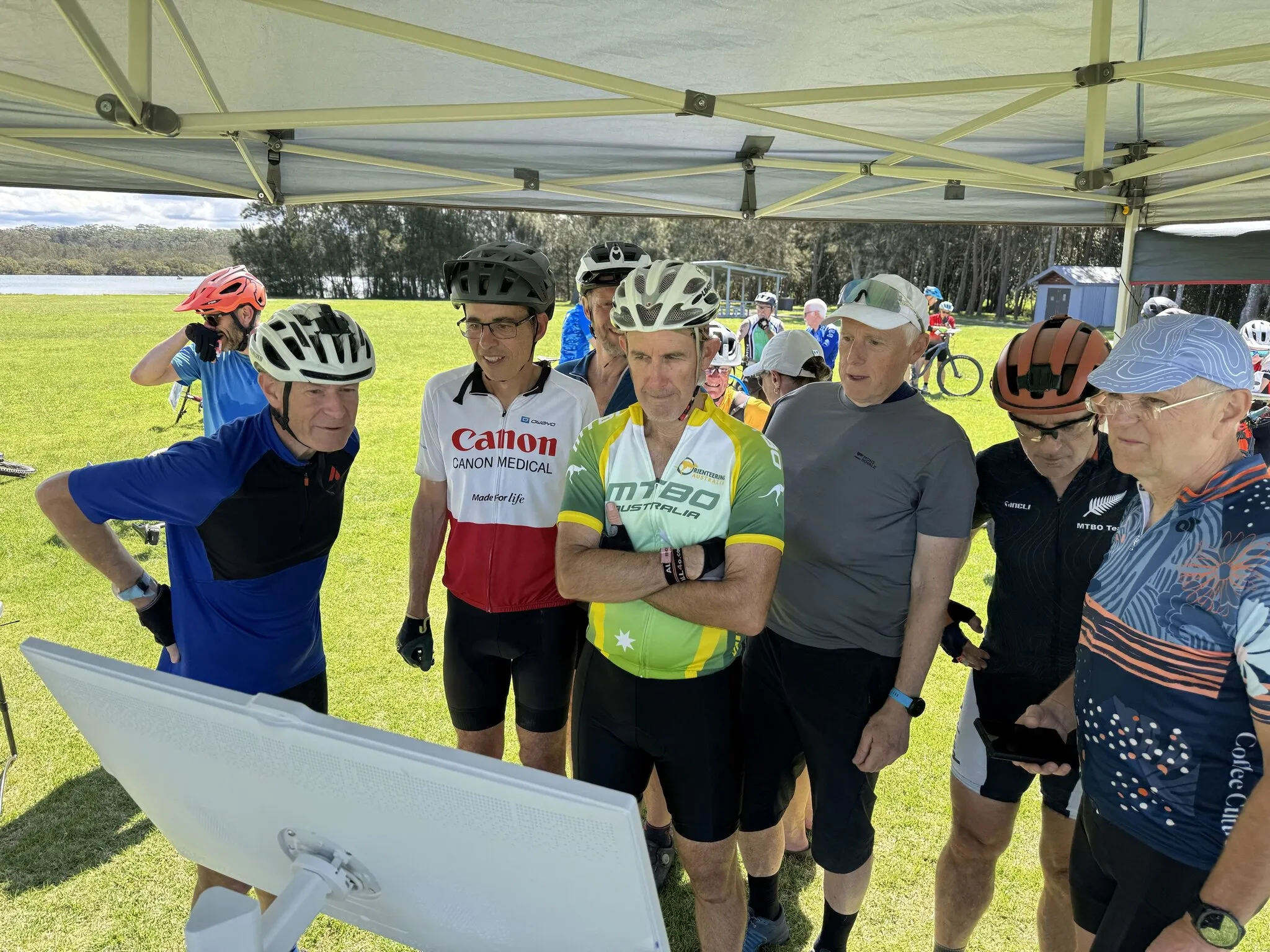 A group of riders in helmets and cycling jerseys crowd round a results screen under a marquee, with grass, trees and water behind them