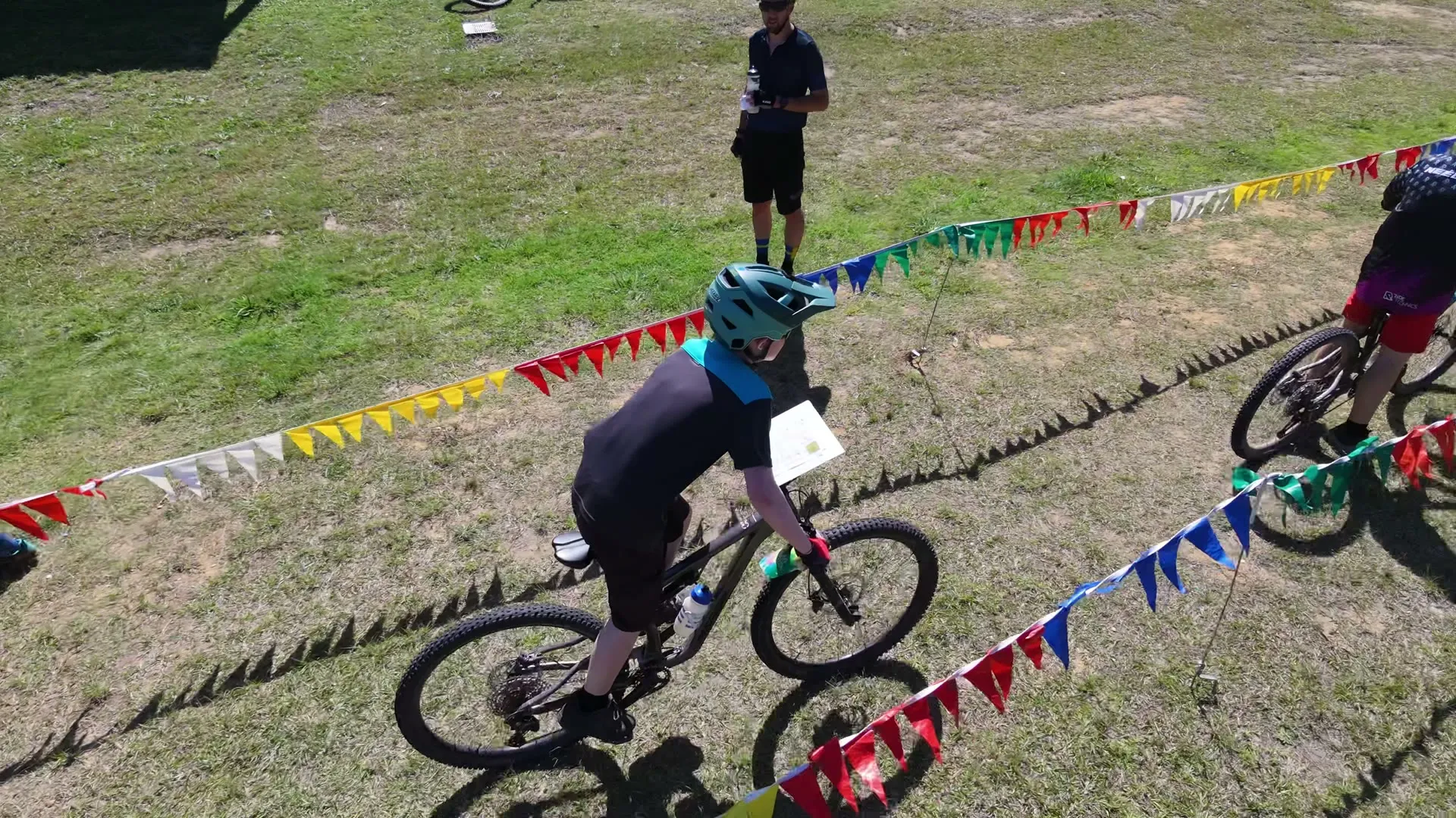 Drone view of two young mountain bike orienteers with maps on their handlebars riding down a finish chute lined with colourful bunting, towards a small finish table on the grass