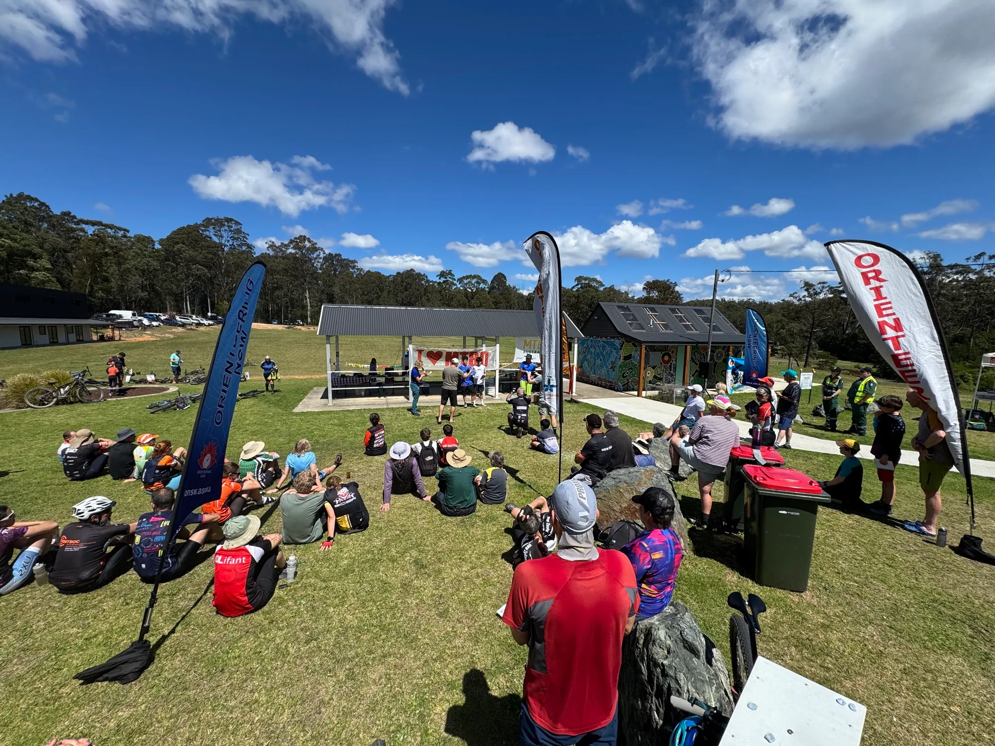 Riders and families sitting on the grass under a blue sky in the Mogo arena, with orienteering flags and bikes around them