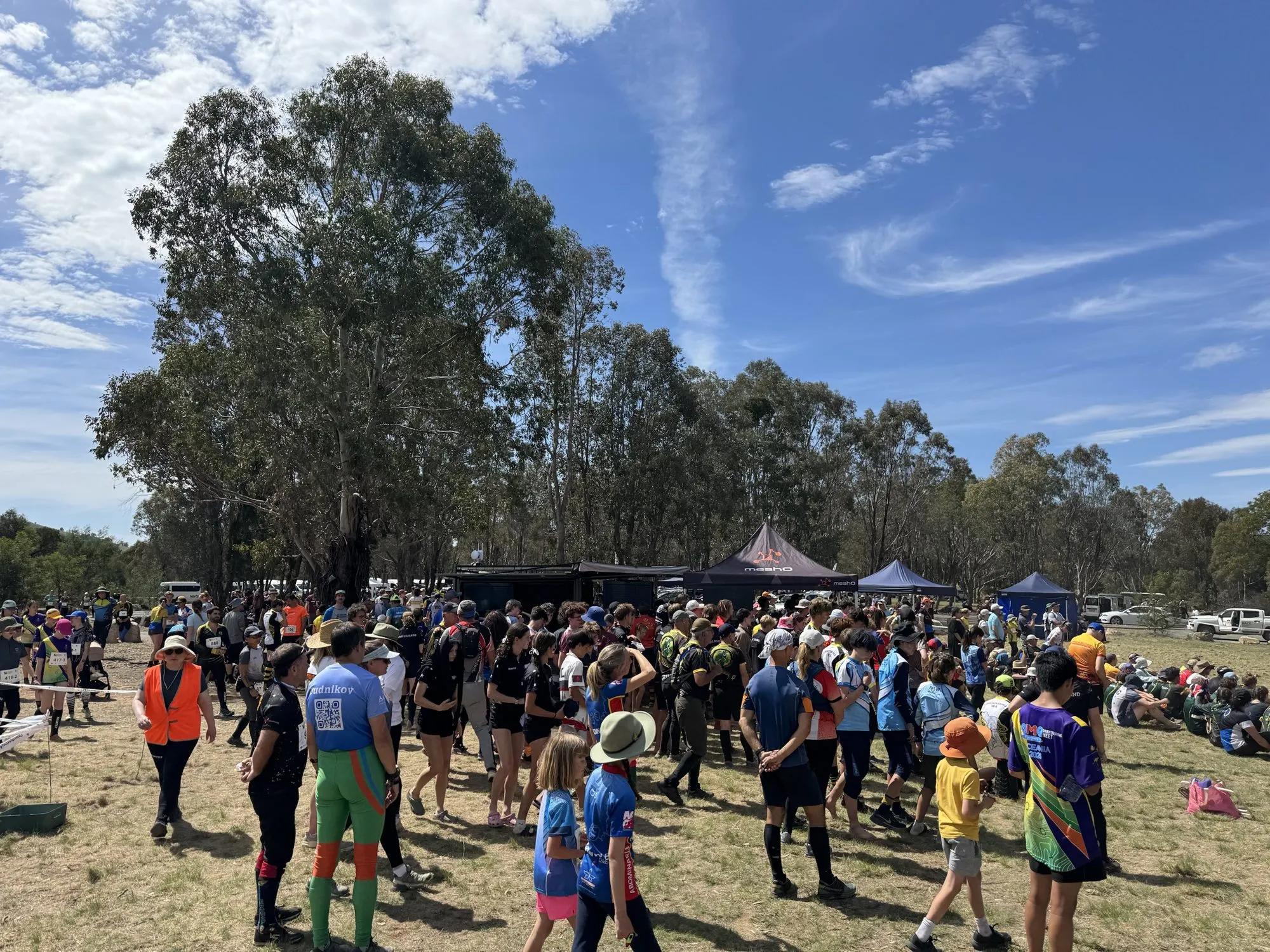 A crowd of competitors in a bush arena under tall gum trees, in front of the meshO results trailer