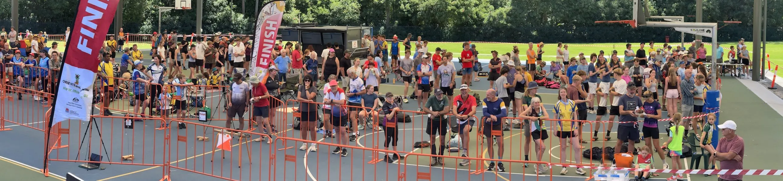 A wide view of the finish arena on a covered sports court, with competitors and spectators lining the orange barriers